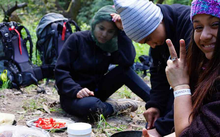 Three outward bound students take a break from backpacking to eat a snack. One of them is giving a peace sign to the camera.